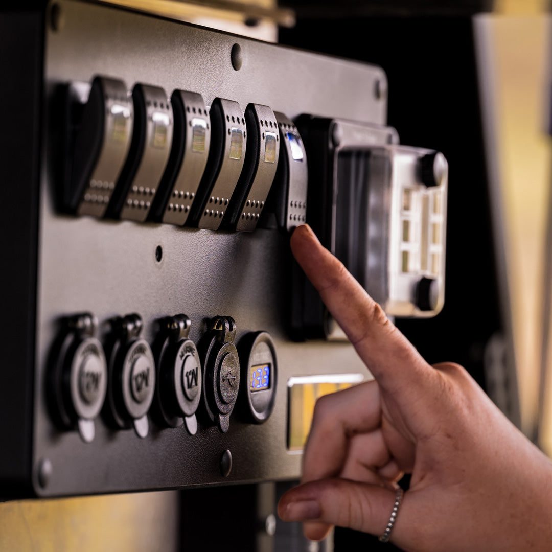 A close-up of a person's hand operating a Mean Mother 12v Power Control Box panel with multiple switches and buttons. The setup includes several labeled switches and electrical sockets, with the hand pressing one of the switches, indicating an industrial or mechanical equipment environment.