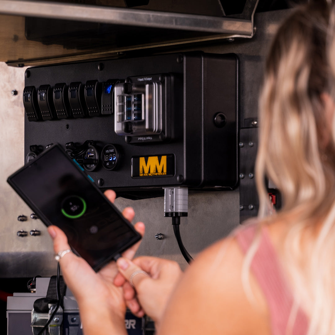 A person with long blonde hair, wearing a pink top, is holding a smartphone near a Mean Mother 12V Power Control Box mounted on a metal surface. The control panel features various switches and a digital display with the "Mean Mother" logo, typically used in canopies and caravans.