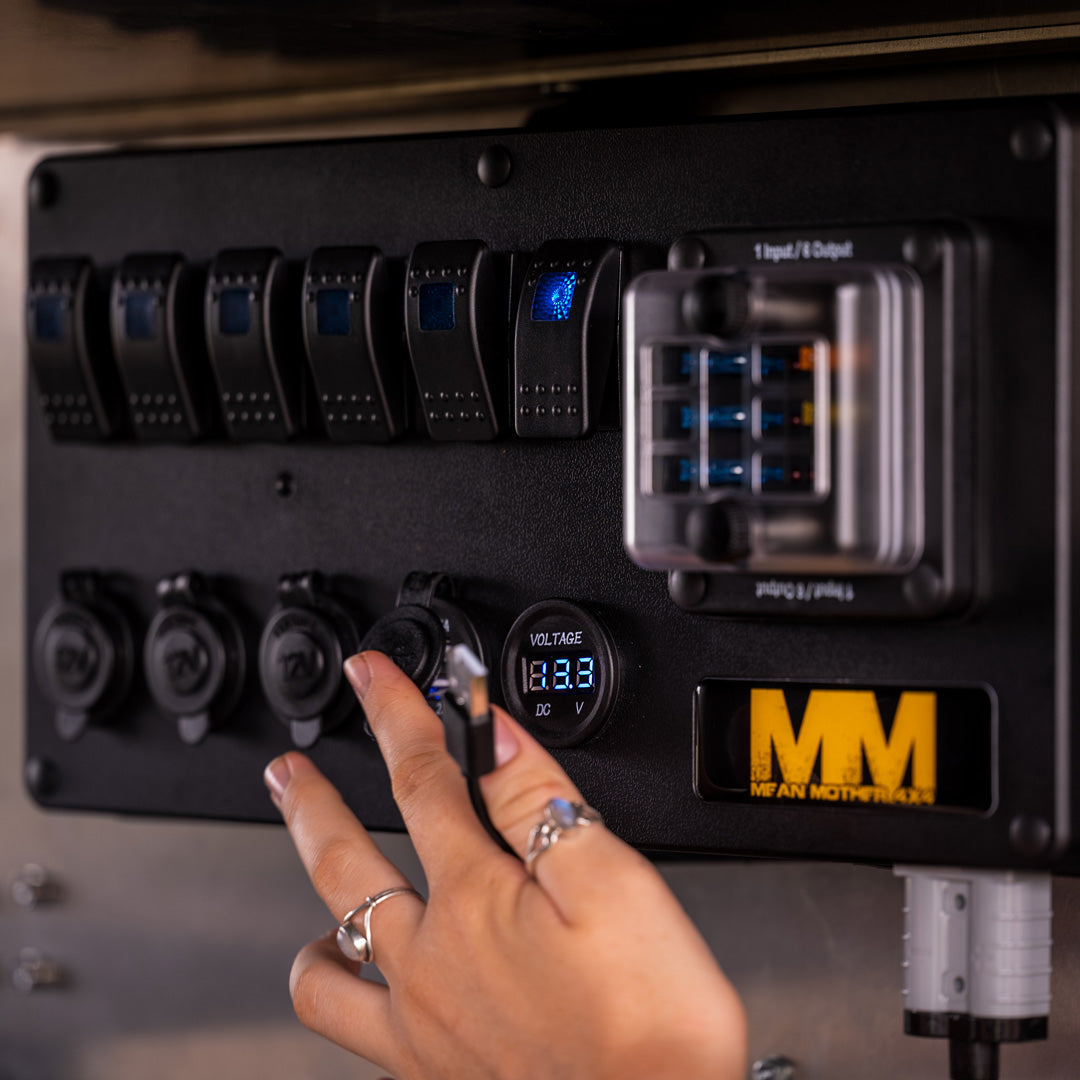 A hand adjusts a knob on the Mean Mother 12v Power Control Box featuring multiple switches, a digital voltage display showing 12.83 volts, and the brand name "Mean Mother" prominently labeled. Rings are visible on the fingers adjusting the knob of this waterproof fuse box.