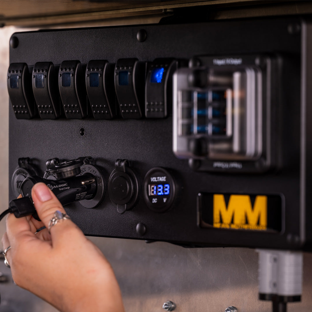 A close-up of a hand plugging a cable into the electrical panel of a Mean Mother 12V Power Control Box. The panel features multiple switches, a voltage meter displaying "13.3V DC," and various connections. The panel includes the Mean Mother logo with "MM" on a black and yellow label. The background is blurred.