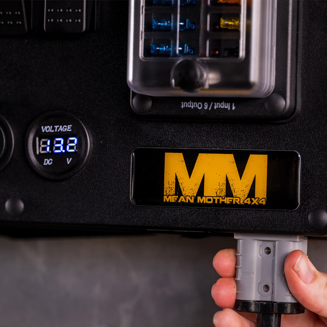 A close-up of the Mean Mother 12v Power Control Box featuring a digital voltage display reading "13.2V" and a waterproof fuse box overhead. The panel showcases the "Mean Mother" logo in bold, distressed yellow font. A hand is seen inserting or removing a plug at the bottom right.