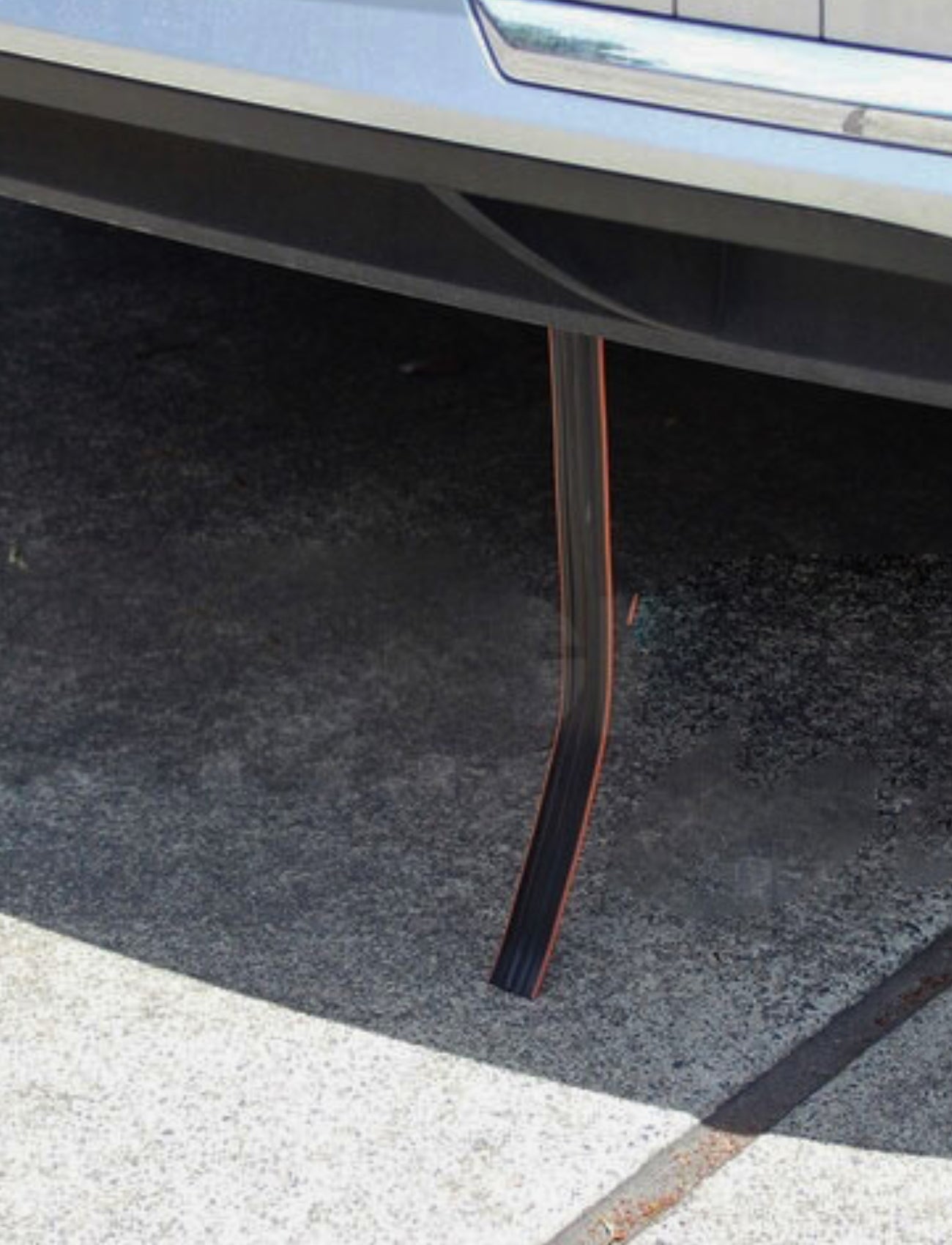 An ANTI STATIC STRAP by Sunland-Protection, in black and flexible design, is partially visible under the silver car bumper, creating a shadow on the pavement. The strap bends towards the ground as if it's a loose piece from the vehicle.