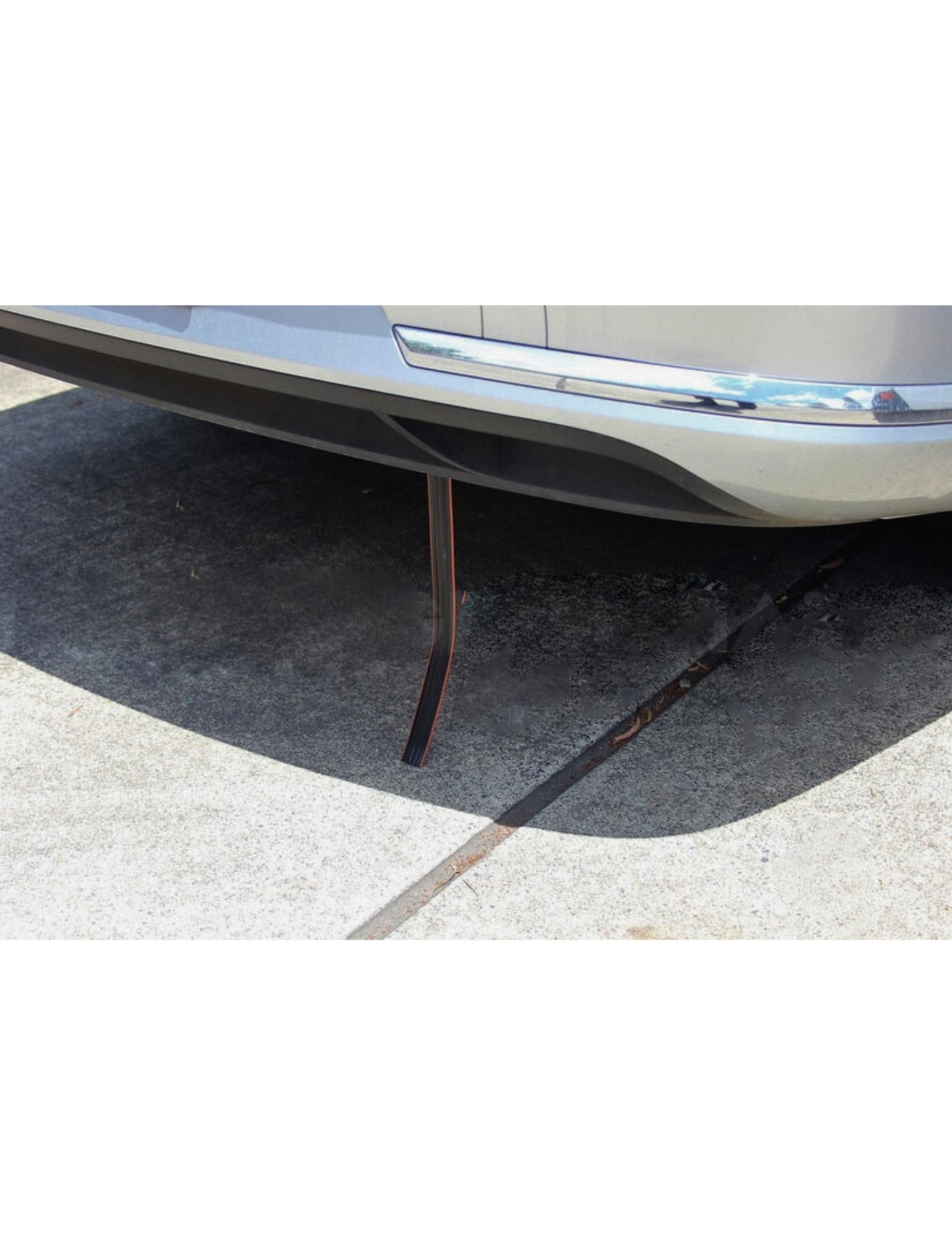A silver car is parked on concrete with a ANTI STATIC STRAP by Sunland-Protection hanging from its rear bumper, designed to safely dissipate static charges with its slightly curved black conductive strap.