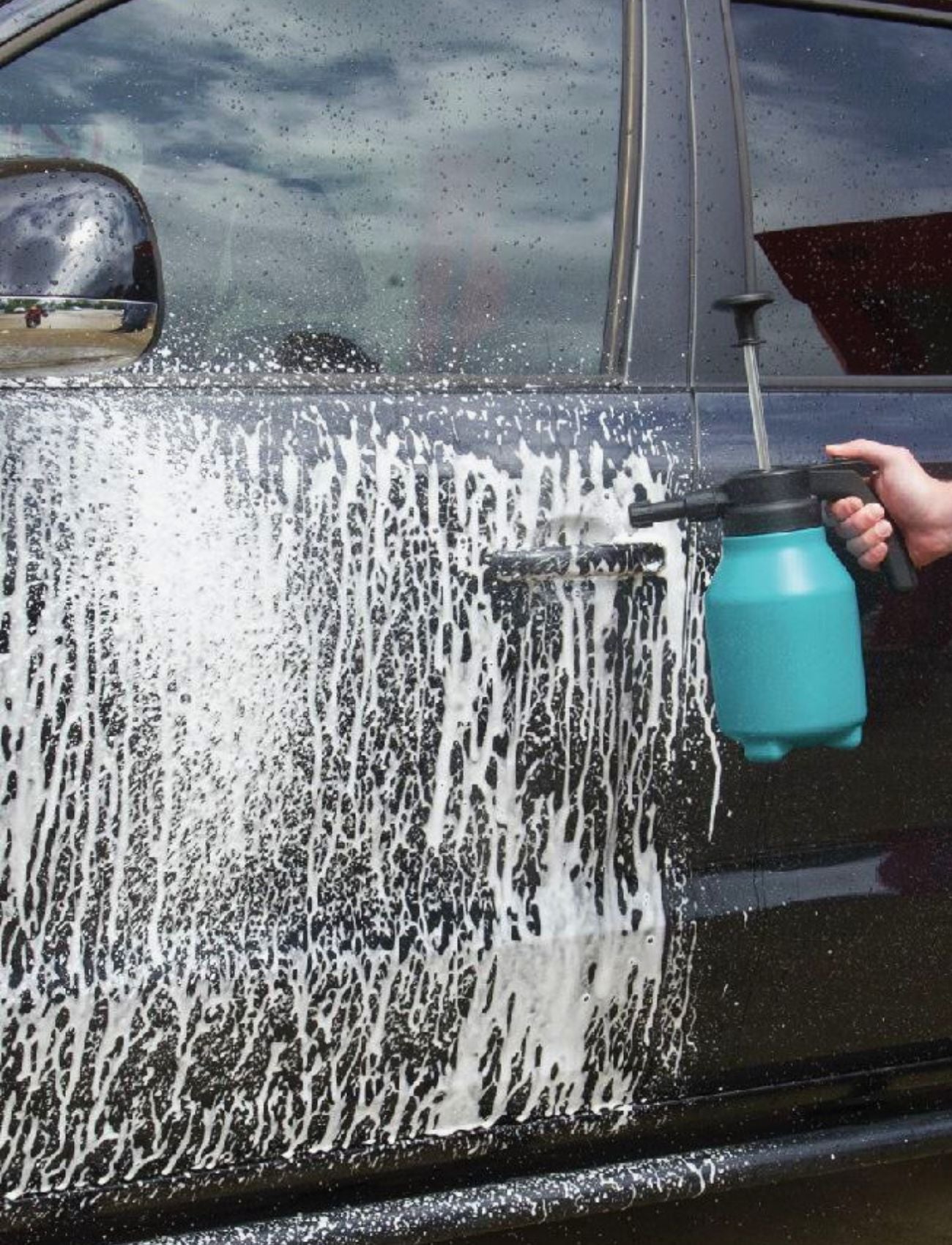 A person uses the Sunland-Protection Foam Pressure Sprayer 2L to generously apply soap to a black car door. Soap drips as they grip the teal bottle with an adjustable spray nozzle, while the car's sleek surface reflects the cloudy sky.