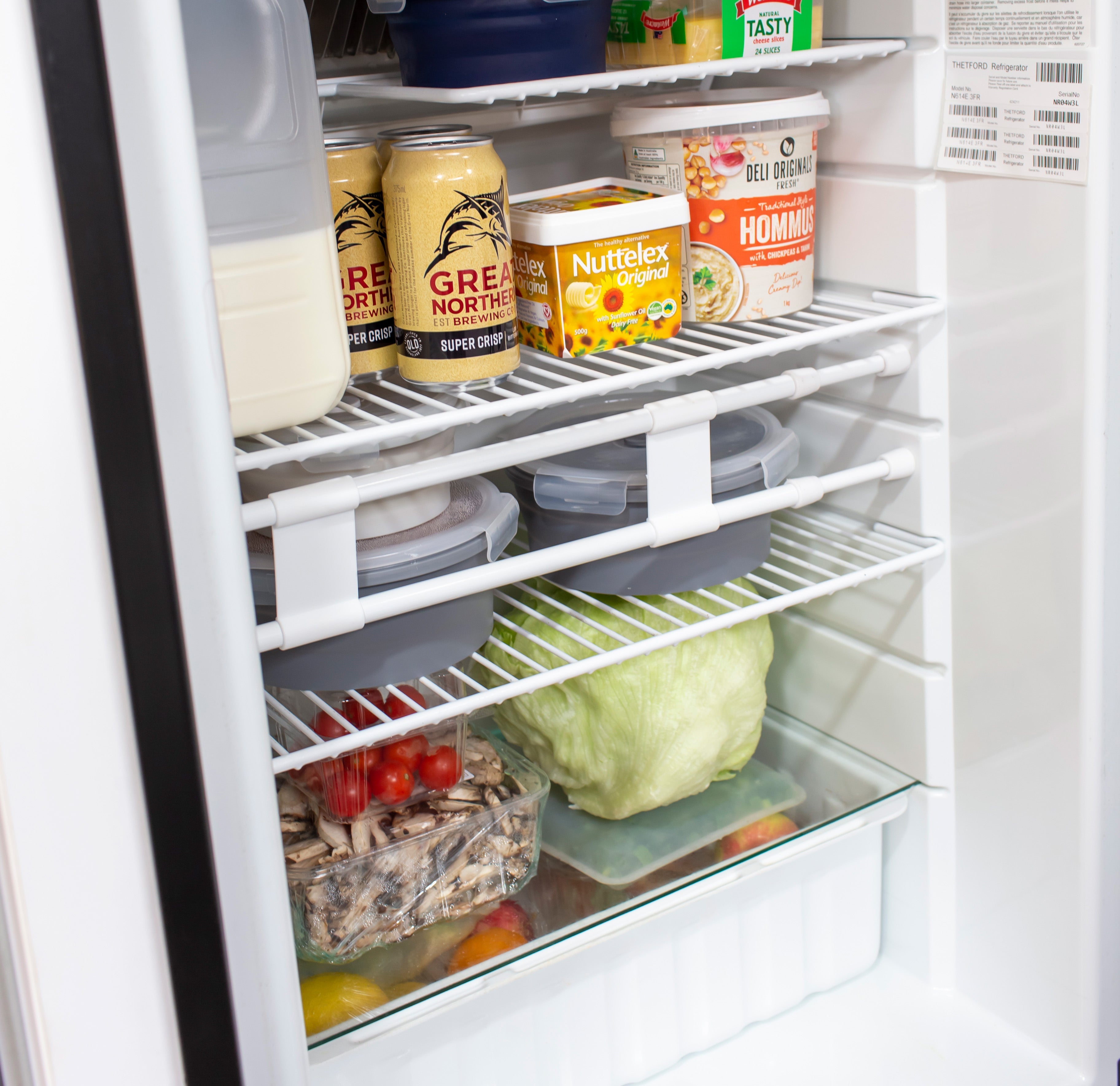 The Explore Caravan Fridge Bars 41-71cm offers an adjustable design with organized shelves, holding two beer cans, a hummus container, a nut spread box, assorted plastic containers, fresh lettuce, tomatoes, mushrooms, and lemons.