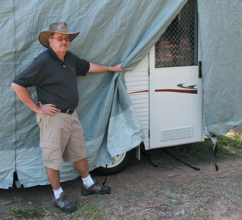 A man in a wide-brimmed hat and sunglasses stands next to a covered trailer, holding a corner of the Prestige Pop Top Caravan Cover 3.97m - 4.27m by Prestige, which partially reveals the trailer's door. His breathable black shirt complements his khaki shorts and dark shoes.