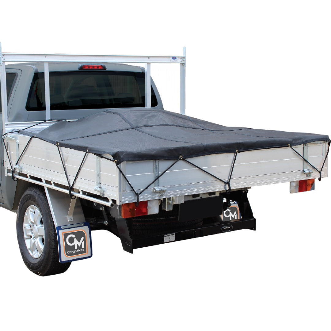 A silver pickup truck is fitted with a flatbed covered by a secured Cargo Mate Trayback Heavy Duty Load Cover - 2x1.8m made of durable Z-Tek mesh material. The flatbed features side panels, and the cover is held in place with bungee cords attached to shock cord fitting points. There is a logo on the mud flaps that reads "CM." The truck is parked on a clear surface.