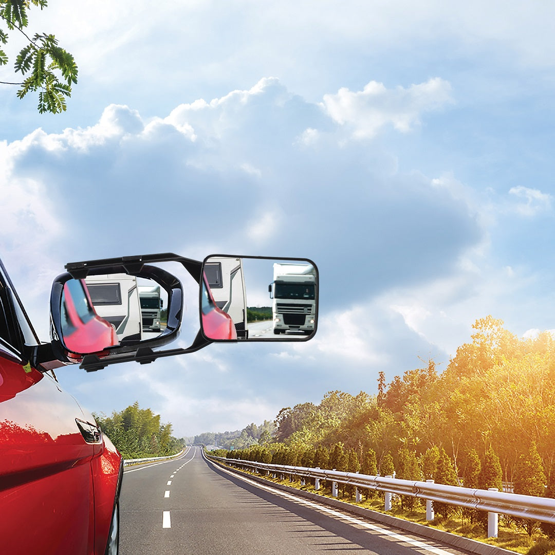 A red vehicle equipped with Drive Multi-Fit Strap-On Towing Mirrors is seen driving on a scenic country road, flanked by trees and bushes, under a blue sky with scattered clouds. The mirrors, known for their easy installation and increased visibility, show the reflection of a large white vehicle following behind.