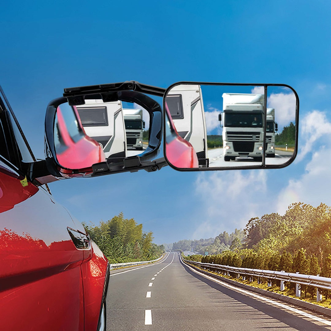 A red car is driving on a clear highway. The side mirror, equipped with a Drive Multi-Fit Strap-On Towing Mirror for increased visibility, reflects a large truck and trailer behind the car. The surrounding landscape features a blue sky, green trees, and roadside barriers.