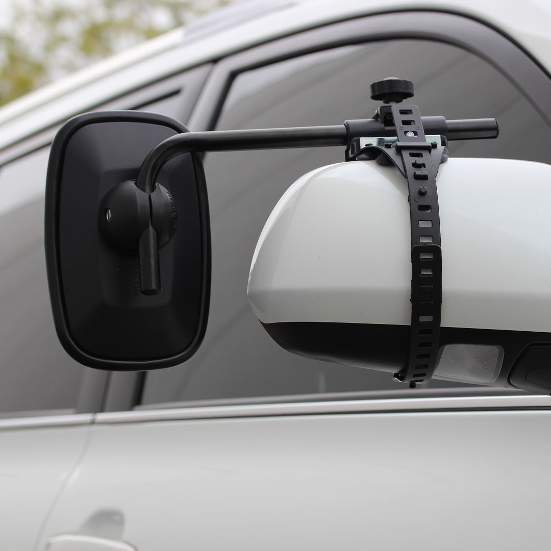 A white vehicle's side mirror includes the Drive brand Towing Mirror, secured with an adjustable black strap for improved rear vision. The vehicle's body and a blurred outdoor scene are visible in the background.