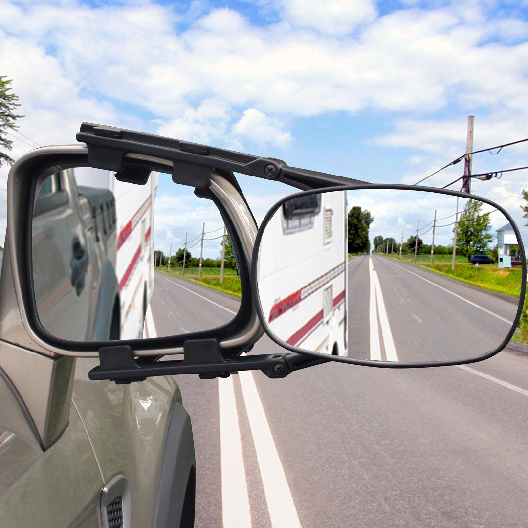Close-up of a vehicle's side mirror with the Drive Towing Mirror Extra Large clipped on. The mirrors reflect a white vehicle with red stripes driving on a rural road lined with trees and power lines under a partly cloudy sky, highlighting the easy installation ideal for travel.