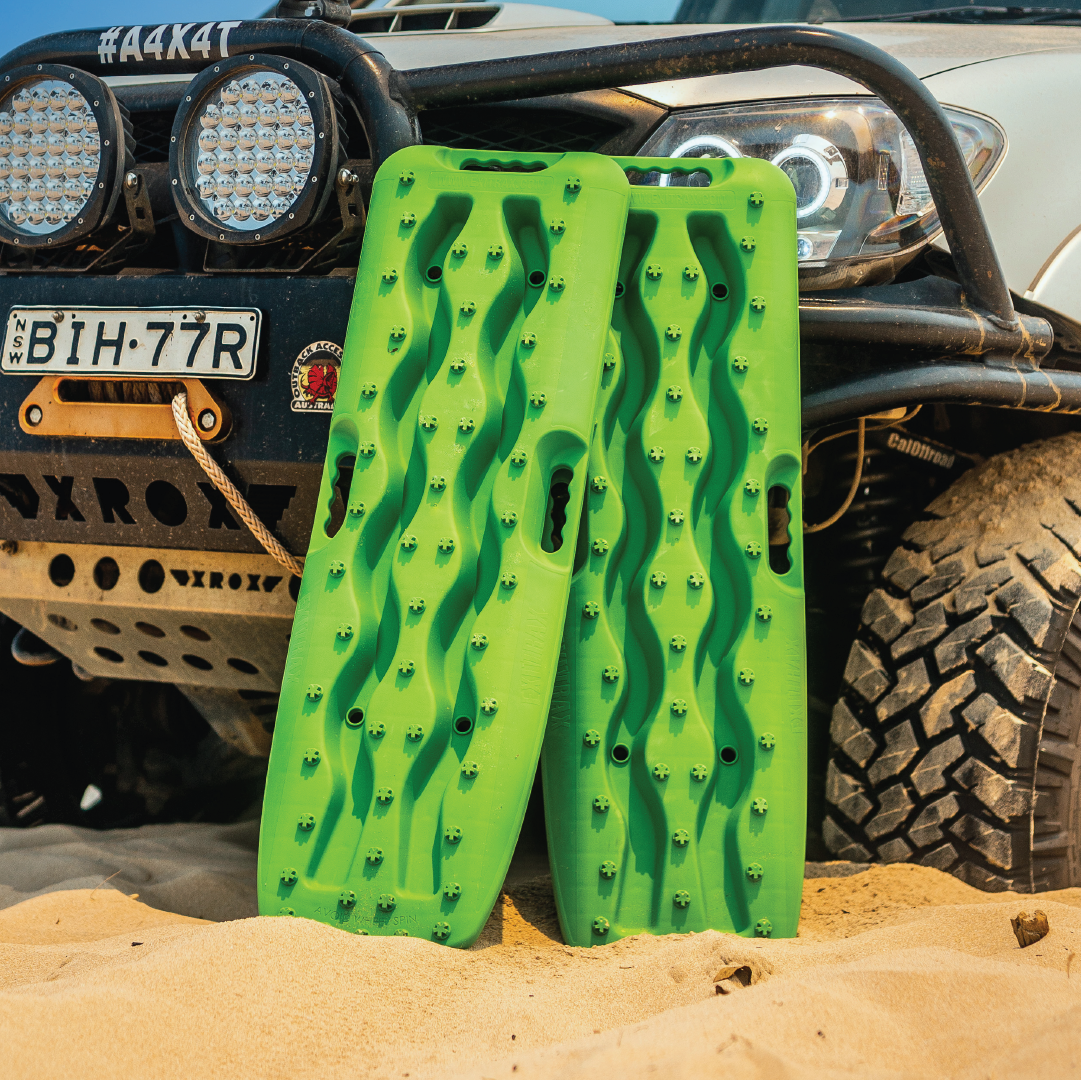 Two bright green Exitrax Recovery Boards 930 Series + Mounts Bundle lean against the front bumper of an off-road vehicle on sandy terrain, with a tire and bull bar seen in the background.