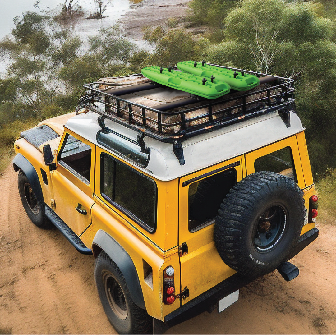 A yellow off-road vehicle with a lifetime warranty traverses a dirt path, outfitted with traction-enhancing Exitrax Recovery Board 930 Series - Black. The spare tire is mounted at the back, and the roof rack is loaded with gear and an inflatable green kayak. Trees and a body of water complete the scenic backdrop.