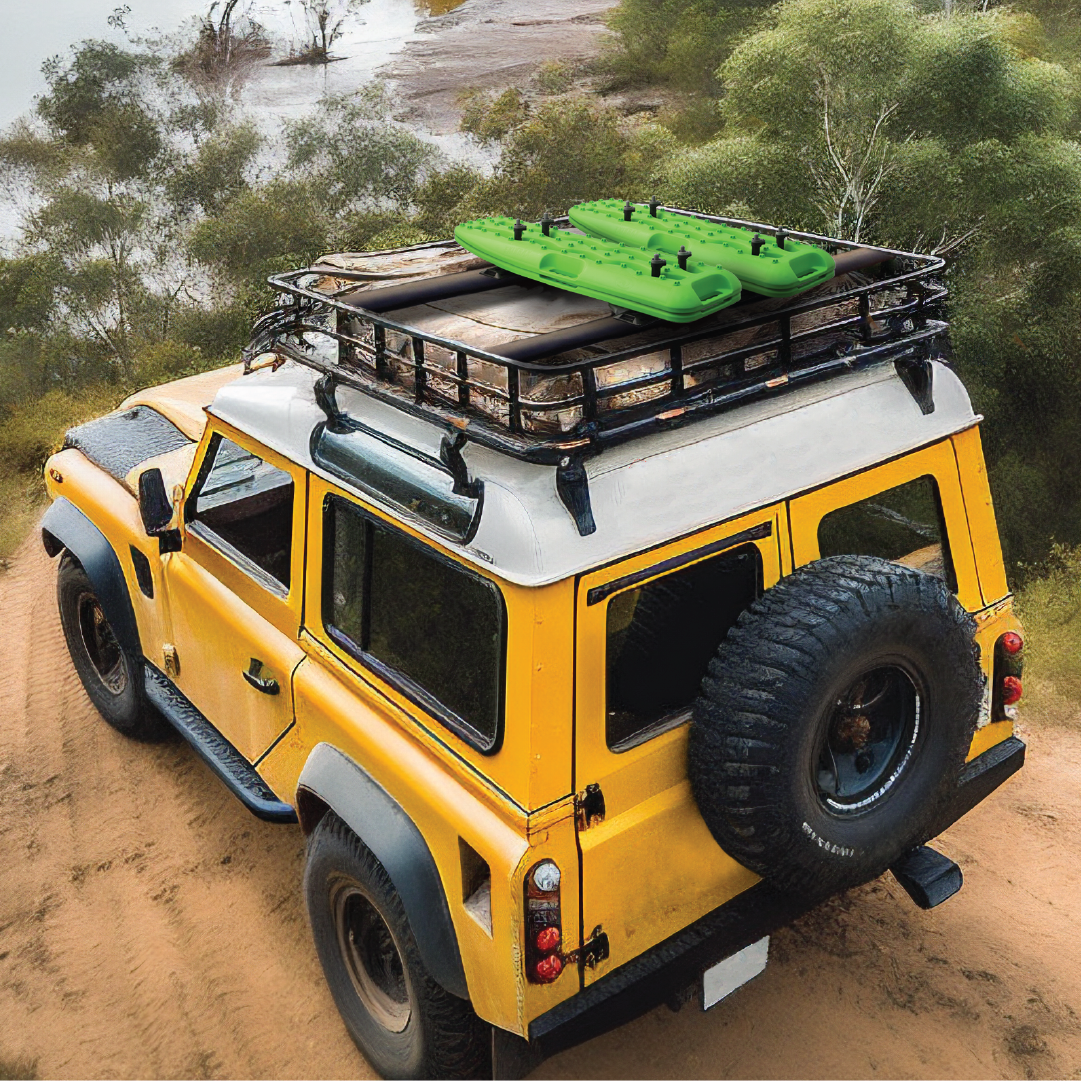 A yellow off-road vehicle travels a dirt path by greenery and water, equipped with a roof rack carrying gear like a green inflatable kayak and featuring Exitrax's Recovery Board 930 Series - Black + Recovery Board Mounts Bundle, along with a spare tire mounted at the back.