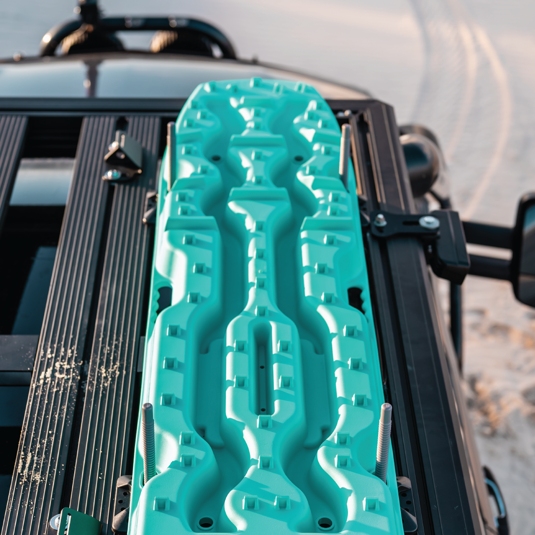 An Exitrax Recovery Board Mounting Kit, featuring a teal recovery board, is installed on the vehicle's roof rack, with sand and tire tracks visible in the background.