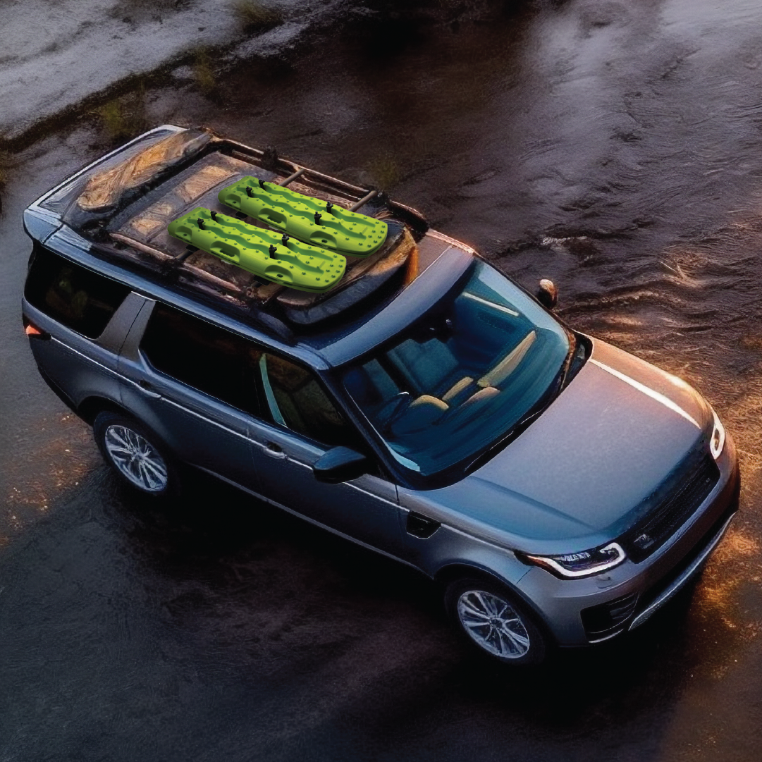 A silver SUV, parked on a wet pavement and viewed from above, showcases bright green inflatable pool floats shaped like cacti fastened on the roof rack with the Exitrax Recovery Board Mounting Kit, known for its corrosion-resistant nylon quick release. The ambient lighting indicates either early morning or evening.