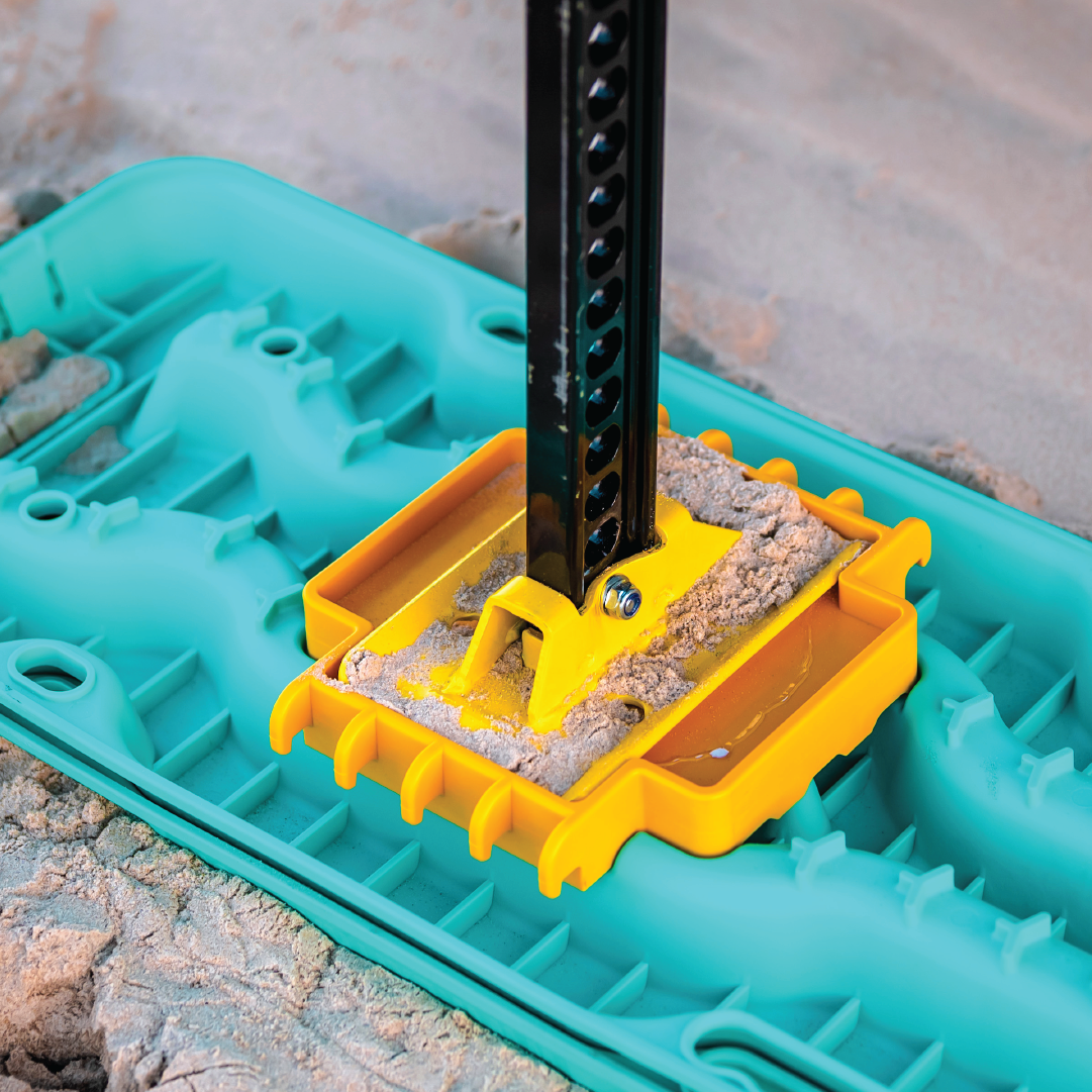 A close-up photo of the Exitrax Recovery Board Link Plate / Jack Base, also known as the Exilink, on sandy ground. The turquoise baseplate, part of the Exitrax Ultimate Series, features a yellow support structure securing a black jack stand. Sand is visible around the baseplate, suggesting equipment setup on a construction or outdoor site.