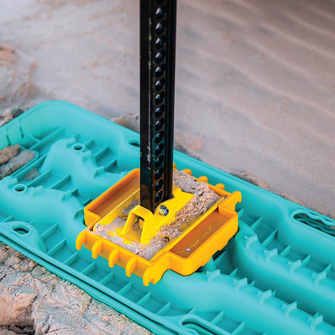 A close-up of a yellow stabilizing plate on soft ground, supporting a jack stand. It sits on an Exitrax Recovery Board Ultimate 1150 in black, which evenly distributes weight to prevent the jack from sinking.