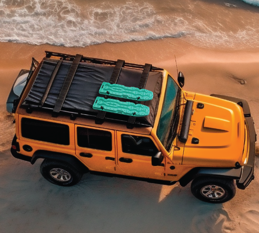 An overhead view of an orange off-road vehicle parked on a sandy beach near the shoreline, with two aqua marine Exitrax Recovery Boards Ultimate 1150 secured on its roof rack. Waves can be seen in the background.