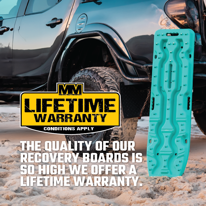 A rugged black off-road vehicle is parked on sandy terrain, with a Blood Orange Exitrax Recovery Board Ultimate 1150 standing upright beside it. A sign indicates, "LIFETIME WARRANTY - Conditions Apply." Our recovery boards provide unmatched quality and traction, ensuring reliable performance for a lifetime.