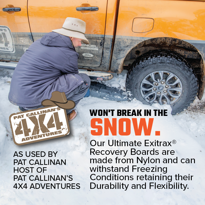 A man in a winter coat kneels to place an Exitrax Recovery Boards Ultimate 1150 - Blood Orange under the tire of a muddy 4x4 on snow, highlighting the bundle’s durability with included Recovery Board Mounts in freezing conditions.
