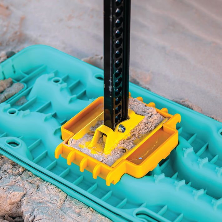 A jack stand is seen on a teal traction mat, secured in a yellow holder and surrounded by sand. Below it lies the Exitrax Recovery Board Ultimate 1150 - Blood Orange with textured grooves for grip, ideal for 4x4 adventures.