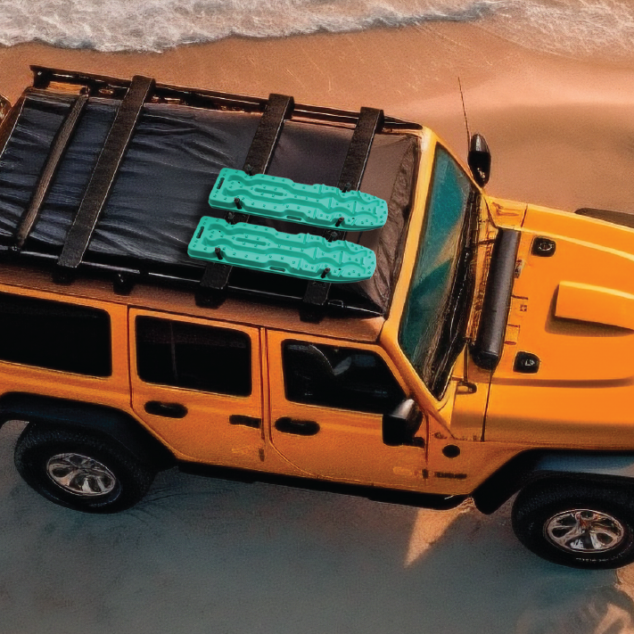 A yellow off-road vehicle is parked on a sandy beach near the waves, with two Exitrax Recovery Boards Ultimate 1150 - Blood Orange and Recovery Board Mounts Bundle secured on the roof rack.