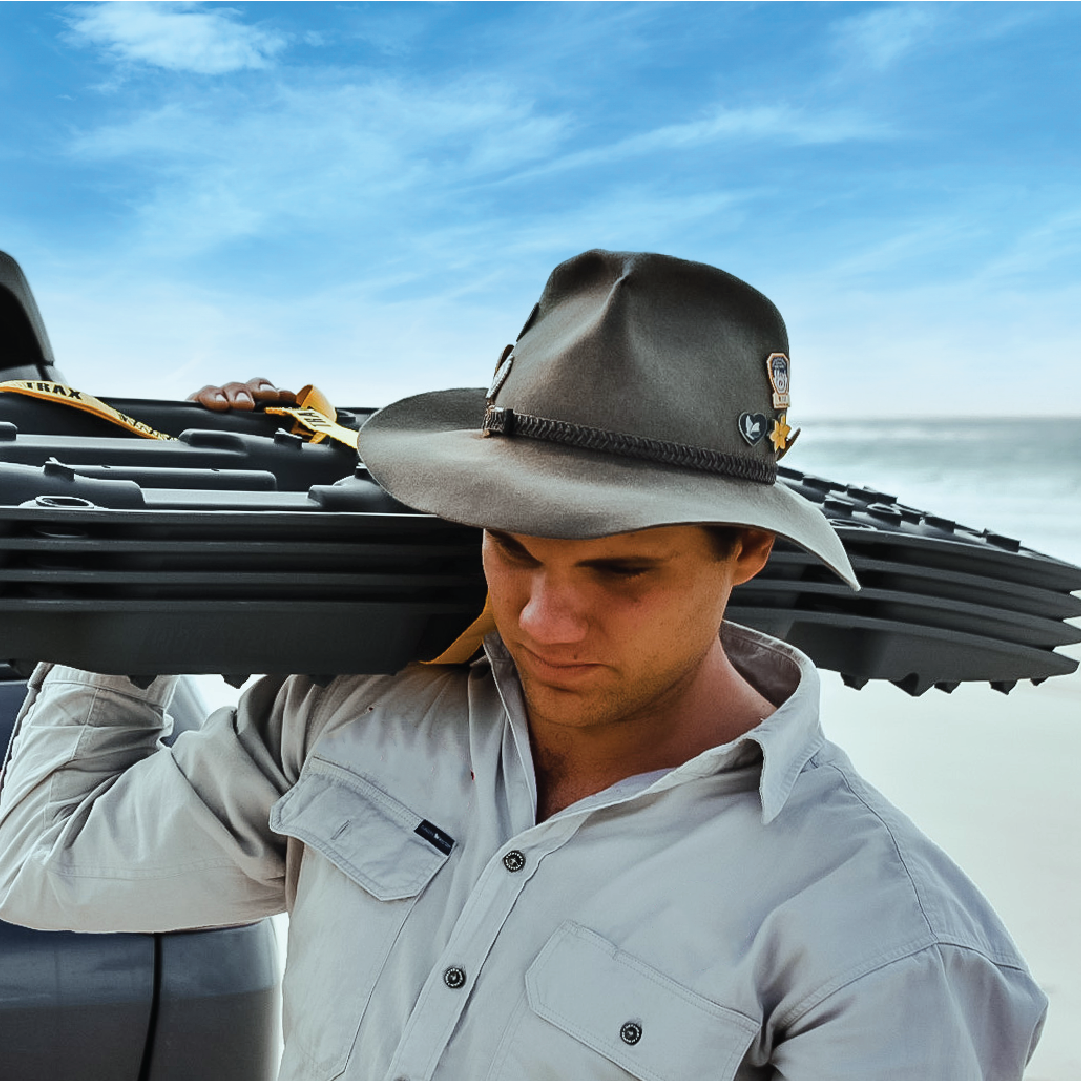 A man in a wide-brimmed hat and light shirt carries an Exitrax Recovery Boards Ultimate 1150 - Gunmetal Grey on his shoulder, standing next to a car on a sandy beach beneath a clear blue sky.