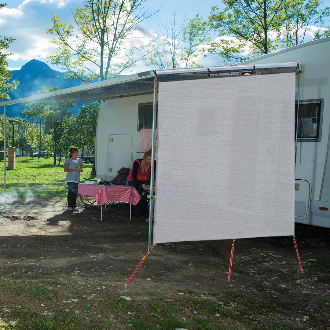 A camping scene with a motorhome parked amidst greenery. An awning extends from the motorhome, equipped with a sunshade and an Explore Caravan End Privacy Screen. A woman sits at a table covered with a pink cloth, while a child stands nearby. Trees and mountains form the backdrop.