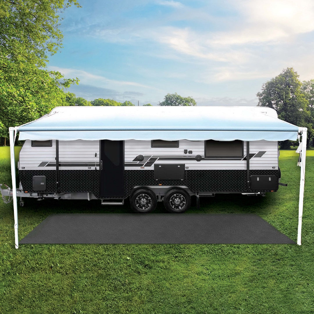 The Explore Caravan & Floor Matting 2.5 x 6m is situated on a grassy field beneath a clear sky, featuring an extended UV-stabilised awning offering shade over its black mat. Lush green trees encircle the scene, forming a tranquil outdoor oasis.