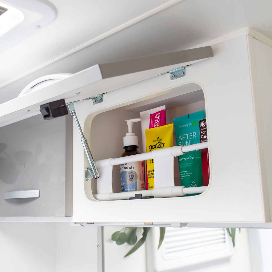 An open bathroom cabinet showcases toiletries such as hand soap, hair gel, aftersun lotion, and face wash. These essentials are neatly organized on two shelves with a sleek white rod featuring an adjustable design reminiscent of Explore's Caravan Fridge Bars (26-43cm) keeping them secure.