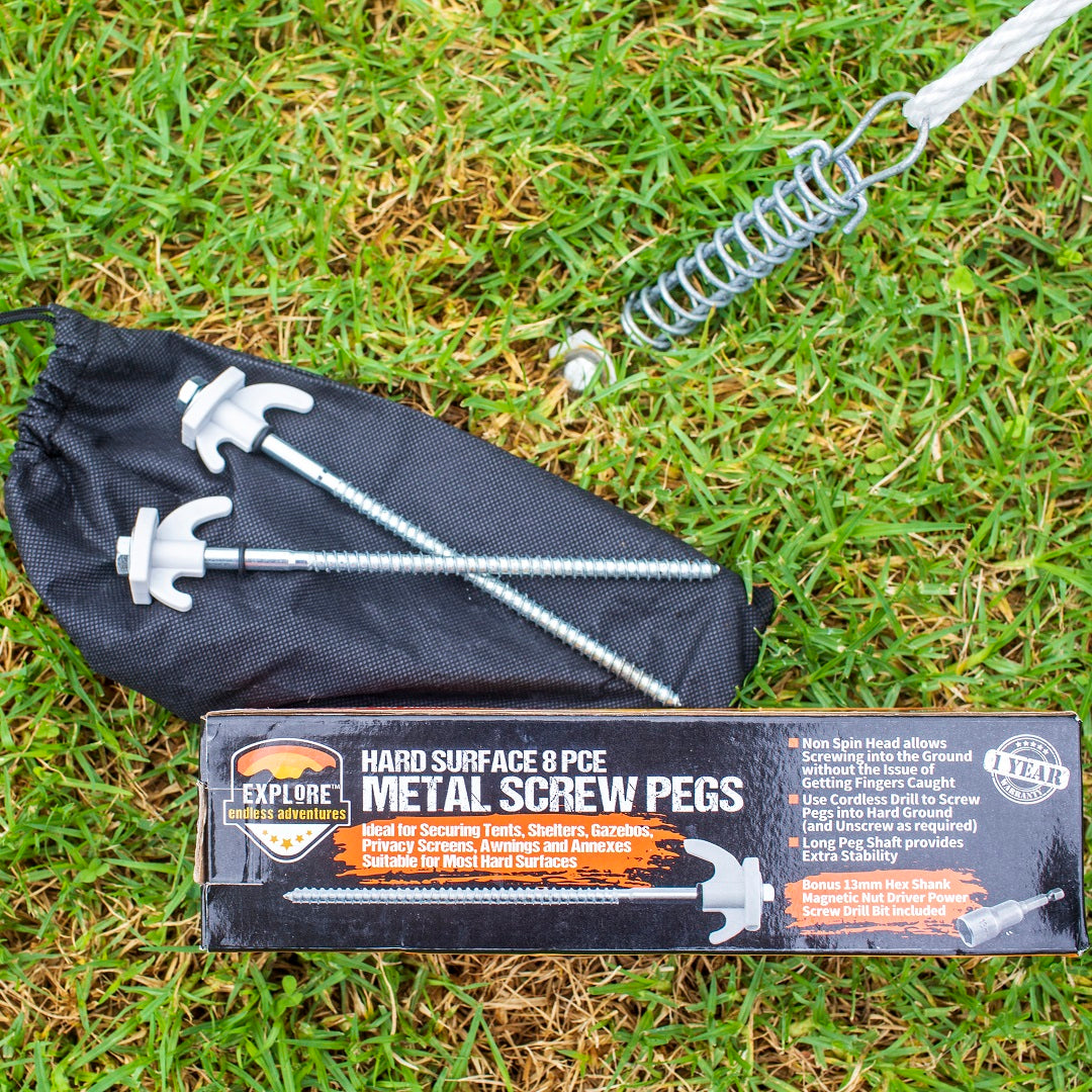 A set of Explore Hard Surface 8 Pce Metal Screw Pegs and a carrying pouch are displayed on grass. The packaging reads "Explore Hard Surface 8 Pce Metal Screw Pegs" and emphasizes their use for securing tents, shades, and more. A coiled spring and cable are also seen alongside the pegs.