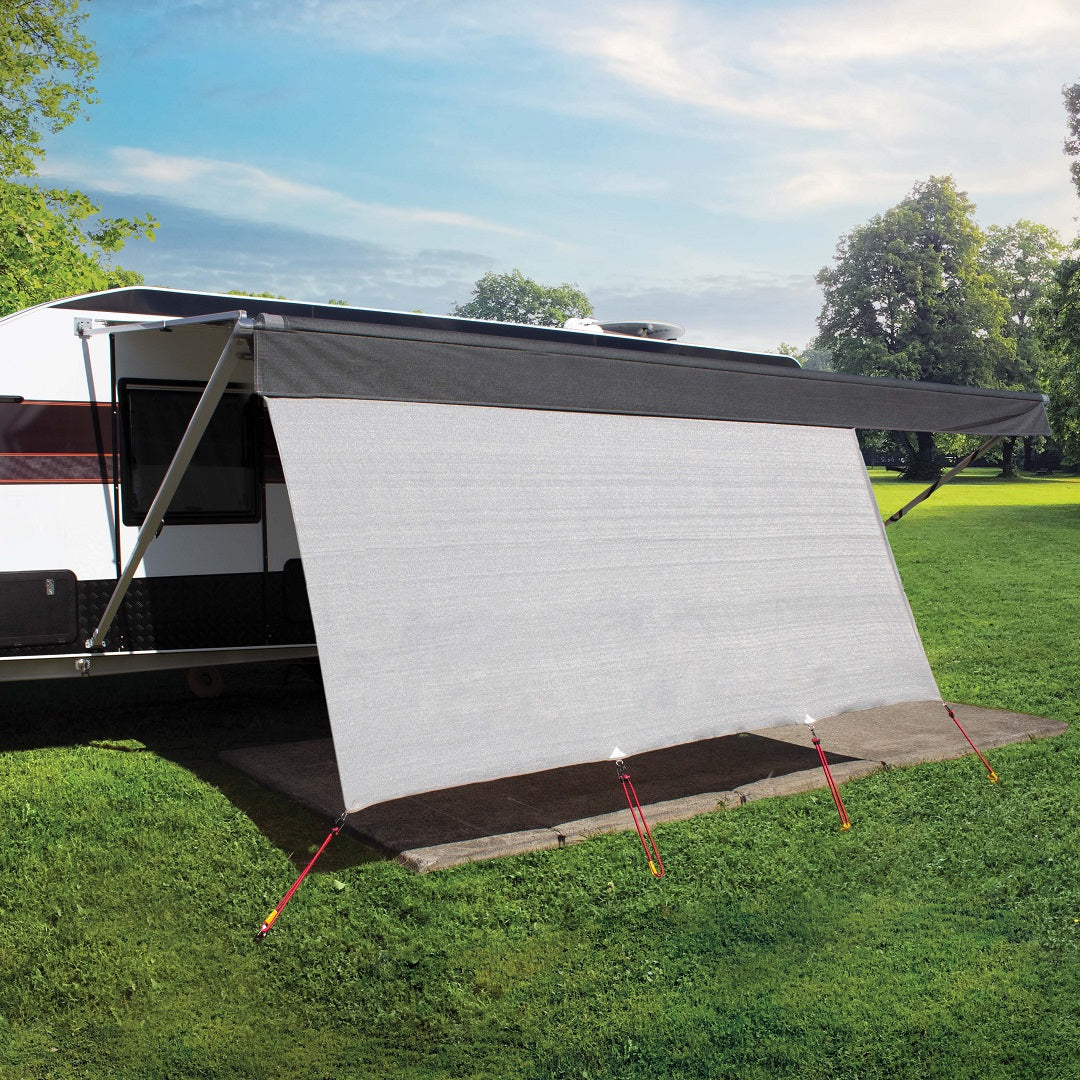 An RV with an Explore Caravan Privacy Screen and extended gray awning is parked on a grassy field surrounded by trees. The awning offers shade, while the breathable mesh screen, secured with red stakes, enhances outdoor living for guests wanting privacy in comfort.