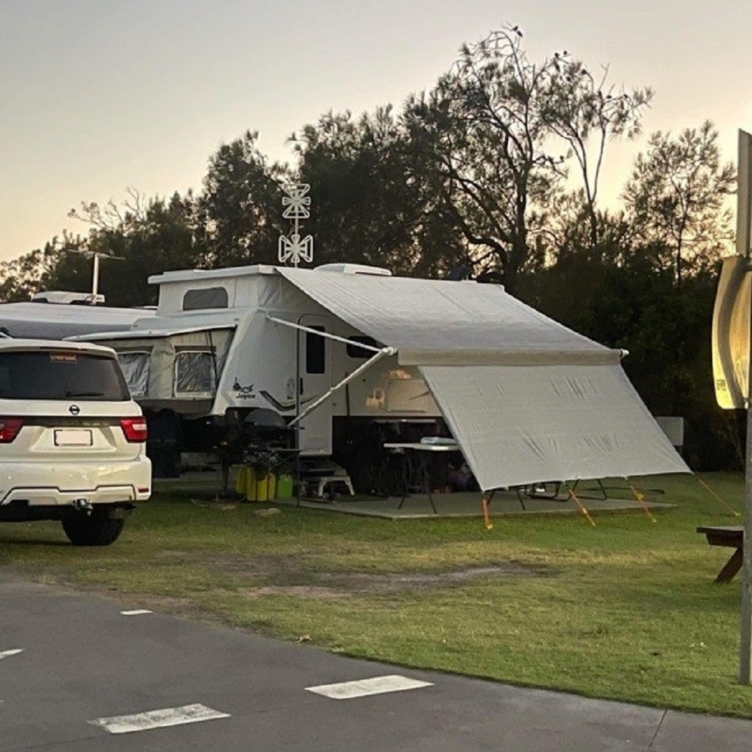An Explore caravan privacy screen (1.8m x 4.3m) enhances a camper trailer with an extended awning on grass, surrounded by trees. A white SUV is parked nearby, camping chairs and a table are set under the awning as the clear evening sky adds to the ambiance.