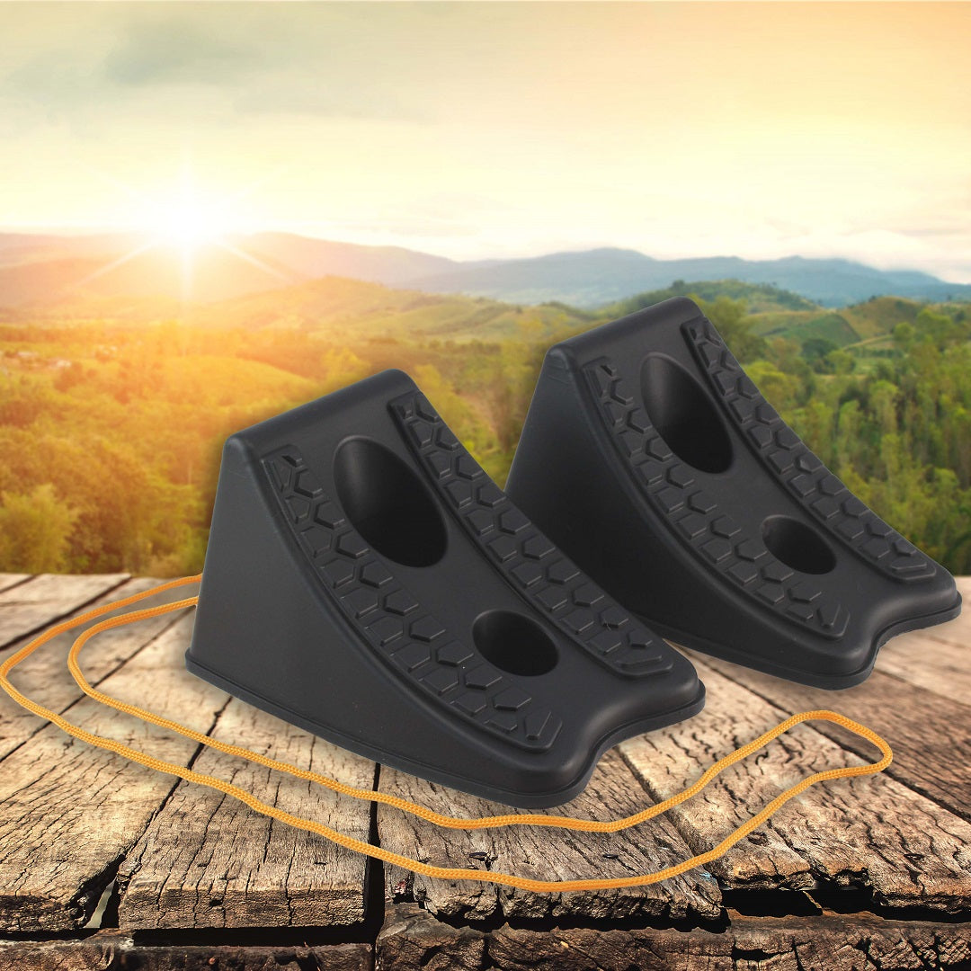 Two Explore Wheel Chocks Nesting - Black with raised textured surfaces are positioned on a wooden surface with a thin rope nearby. Behind them, the scenic view showcases rolling green hills under a sunny sky.