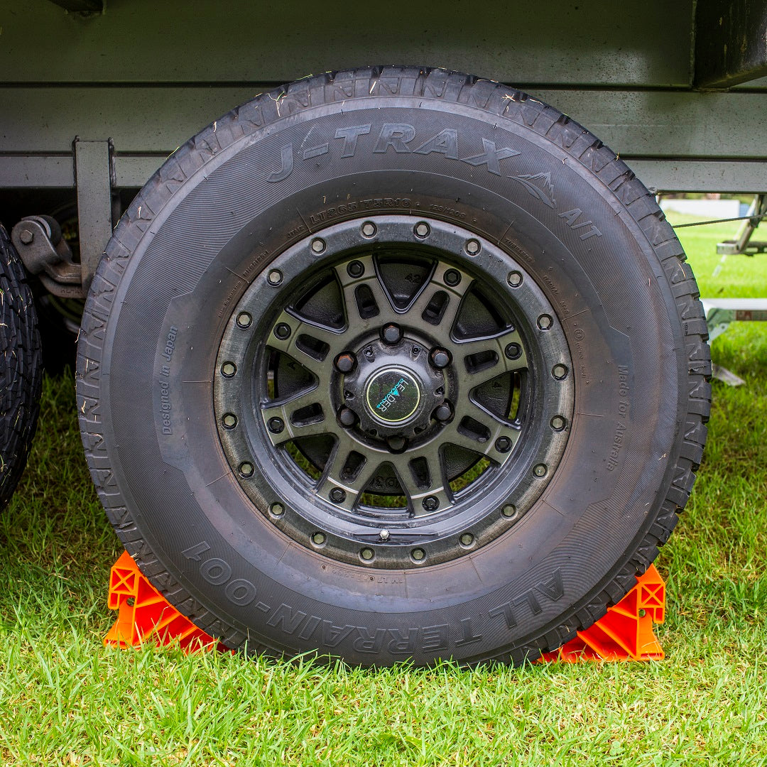 A close-up of a large tire with a rugged tread pattern is supported by Explore Wheel Chocks, which are bright orange for high visibility. Made in Australia, the tire rests on a grassy surface, ensuring the parked vehicle's stability.