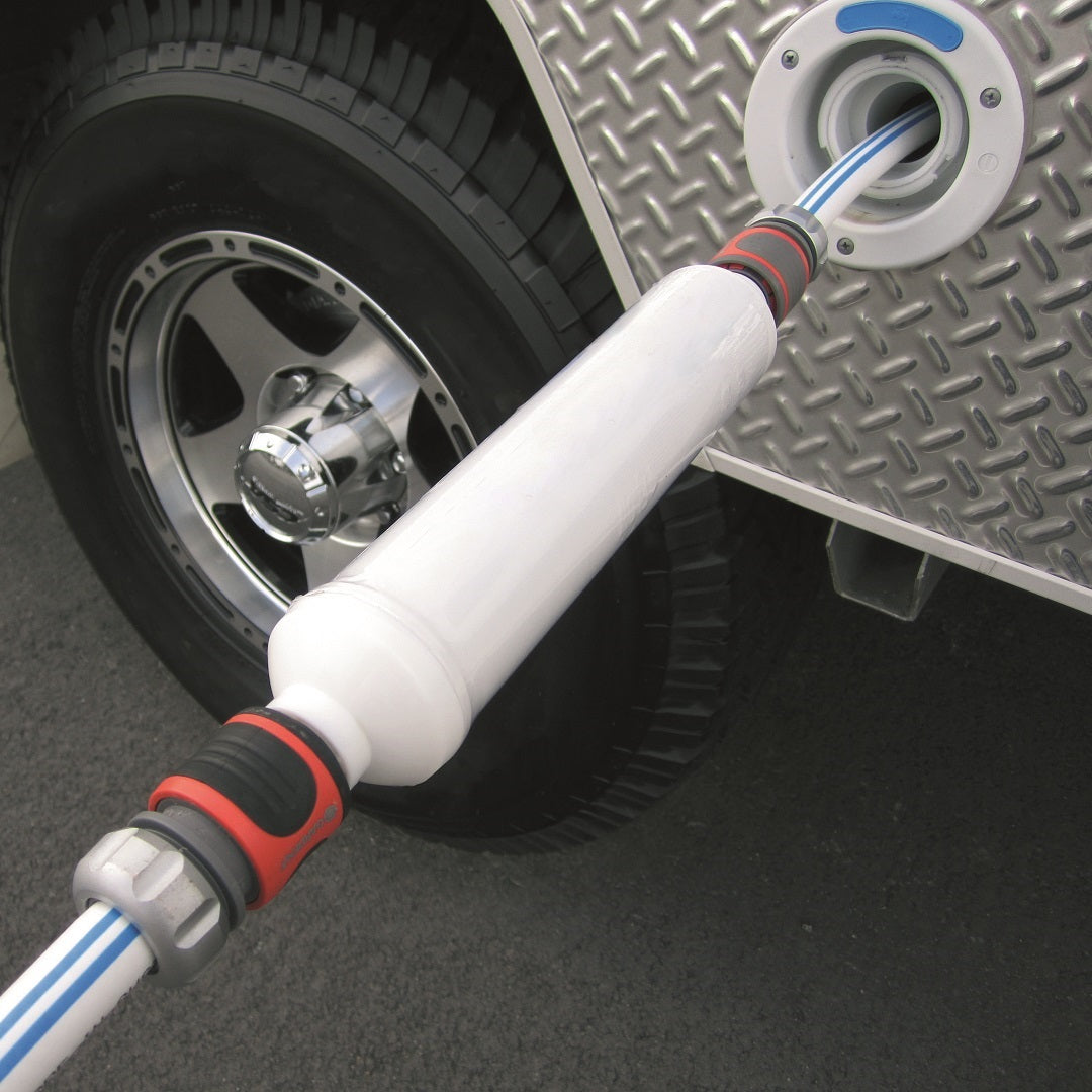 Close-up view of a fresh water hose with an Explore Inline Water Filter Housing connected to an RV's water inlet. The background shows part of the RV's diamond-plated exterior and a portion of a tire with a metallic rim. The hose has red, black, and white fittings, ensuring safe drinking water on the go.