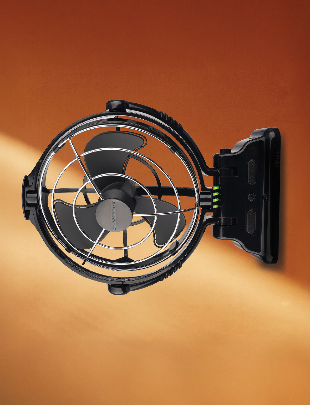 The Explore Caravan Fan Black | Carabreeze, a wall-mounted, whisper-quiet three-blade fan with a metal safety grille, is set against a warm orange background with light streaming in from the left.
