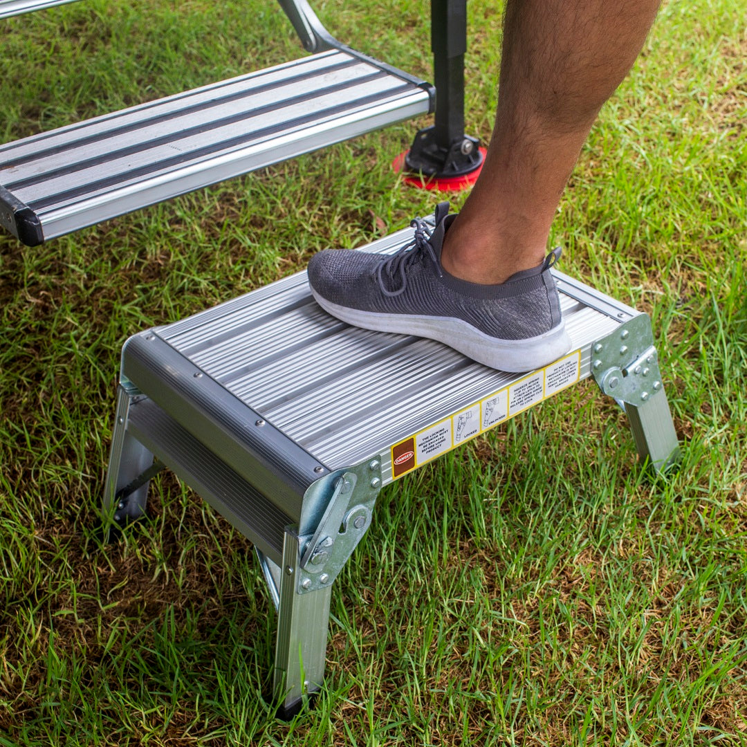 A person wearing gray sneakers steps onto the lightweight Explore Folding Step Stool - Aluminum, set on green grass. The stool has a ribbed surface, non-slip feet, and warning labels on the side. A larger set of steps is partially visible in the background.