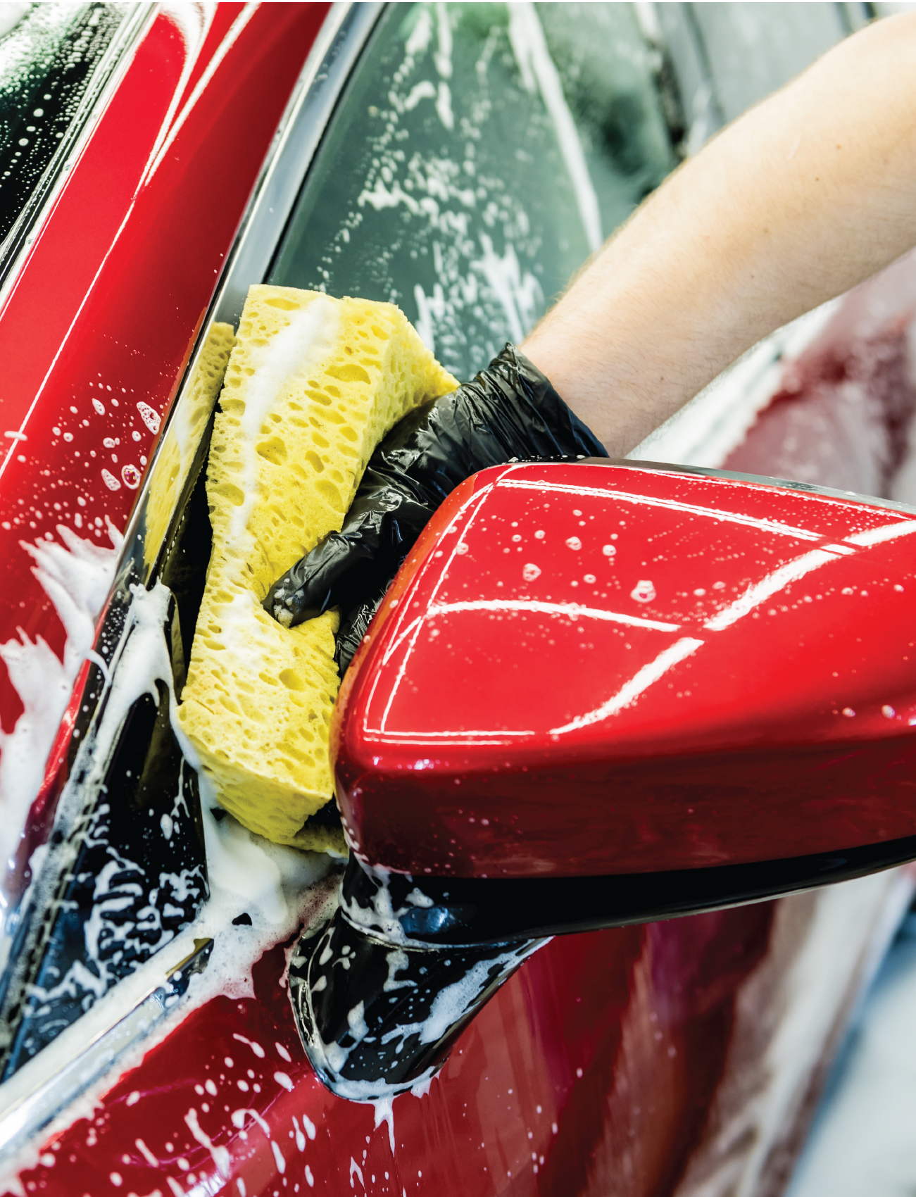 A person in a black glove uses the durable SPONGE ALL PURPOSE (GIANT SIZE) by Sunland-Protection to wash a red car. The high sudsing action covers parts of the vehicle as the yellow sponge cleans near the side mirror.
