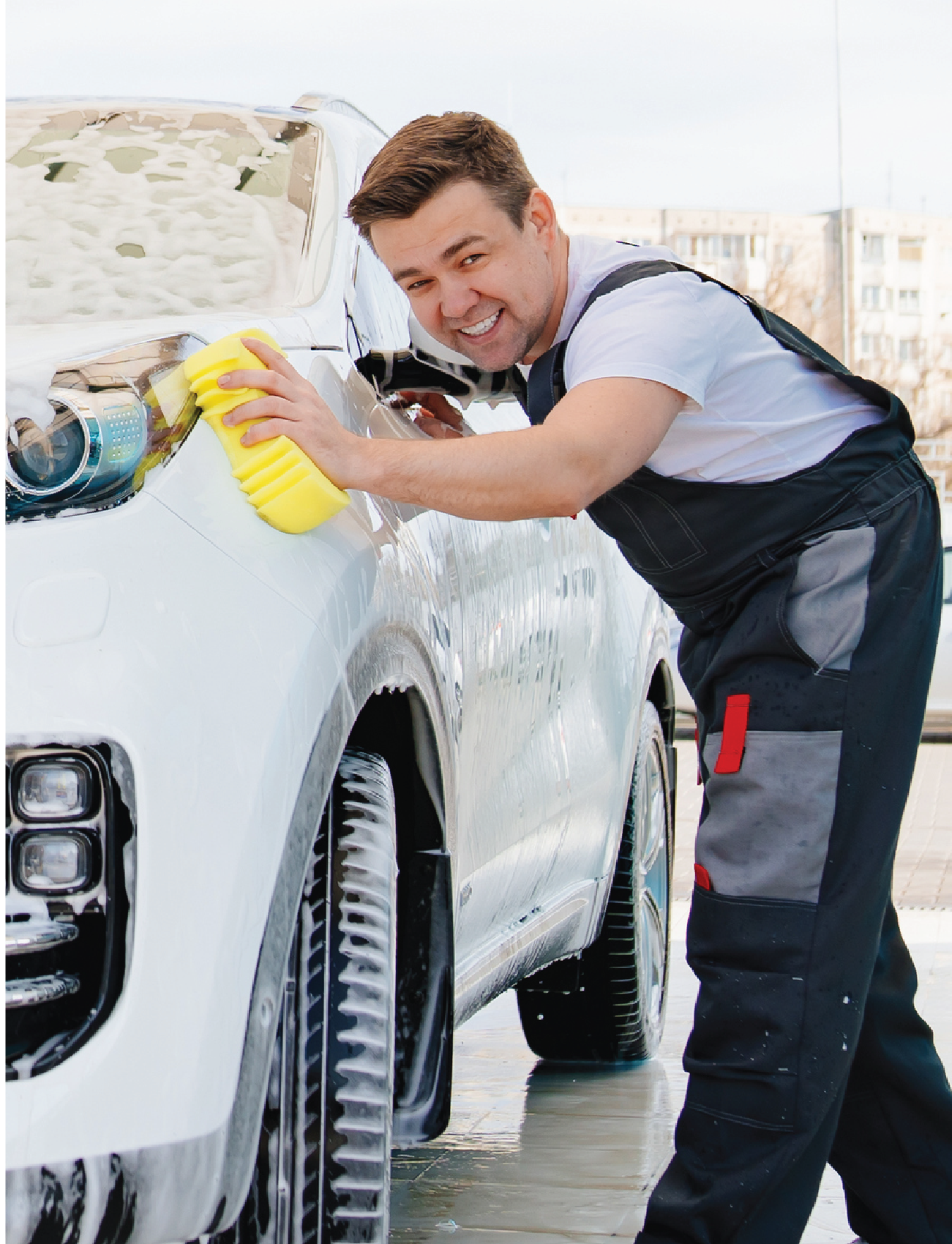 A man in a white shirt and overalls smiles as he washes a white car with Sunland-Protection's SPONGE ALL PURPOSE (LARGE SIZE). The car is covered in soap suds due to the high sudsing action, and the scene is set outdoors in a city area.