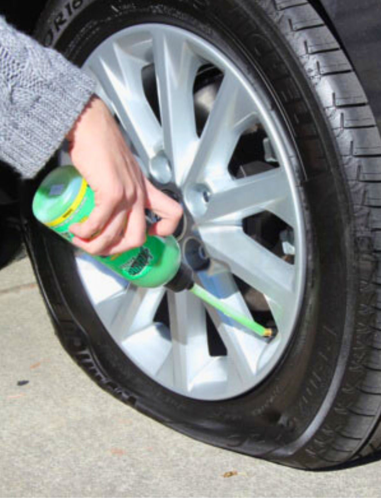 Wearing a gray sweater, someone uses a green bottle of Liquid Patch 20L tyre sealant to fix a flat car tire on a paved surface. Sunlight highlights the scene as they efficiently repair the puncture.