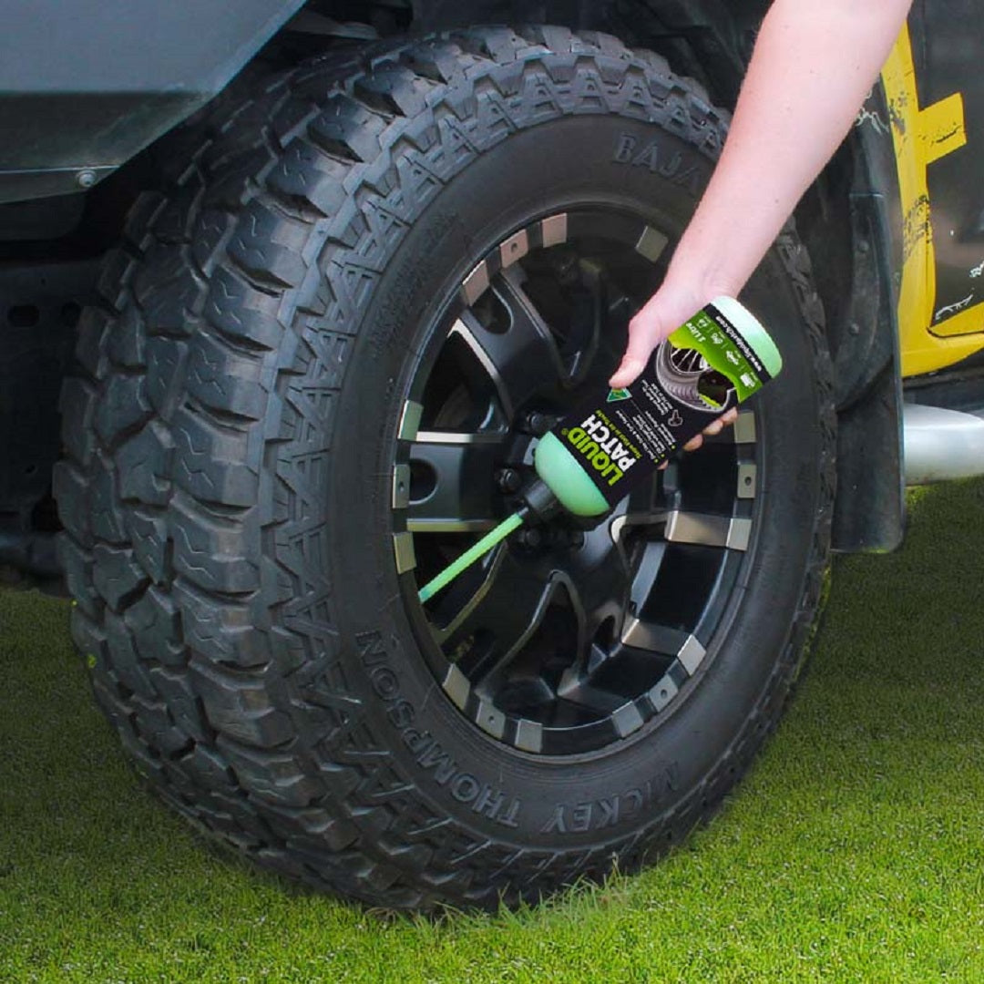 An individual applies Liquid Patch Tyre Puncture Repair - 250ml to the tire of a black vehicle parked on grass. The tire, which provides puncture protection, showcases bold tread and features a five-spoke rim design.