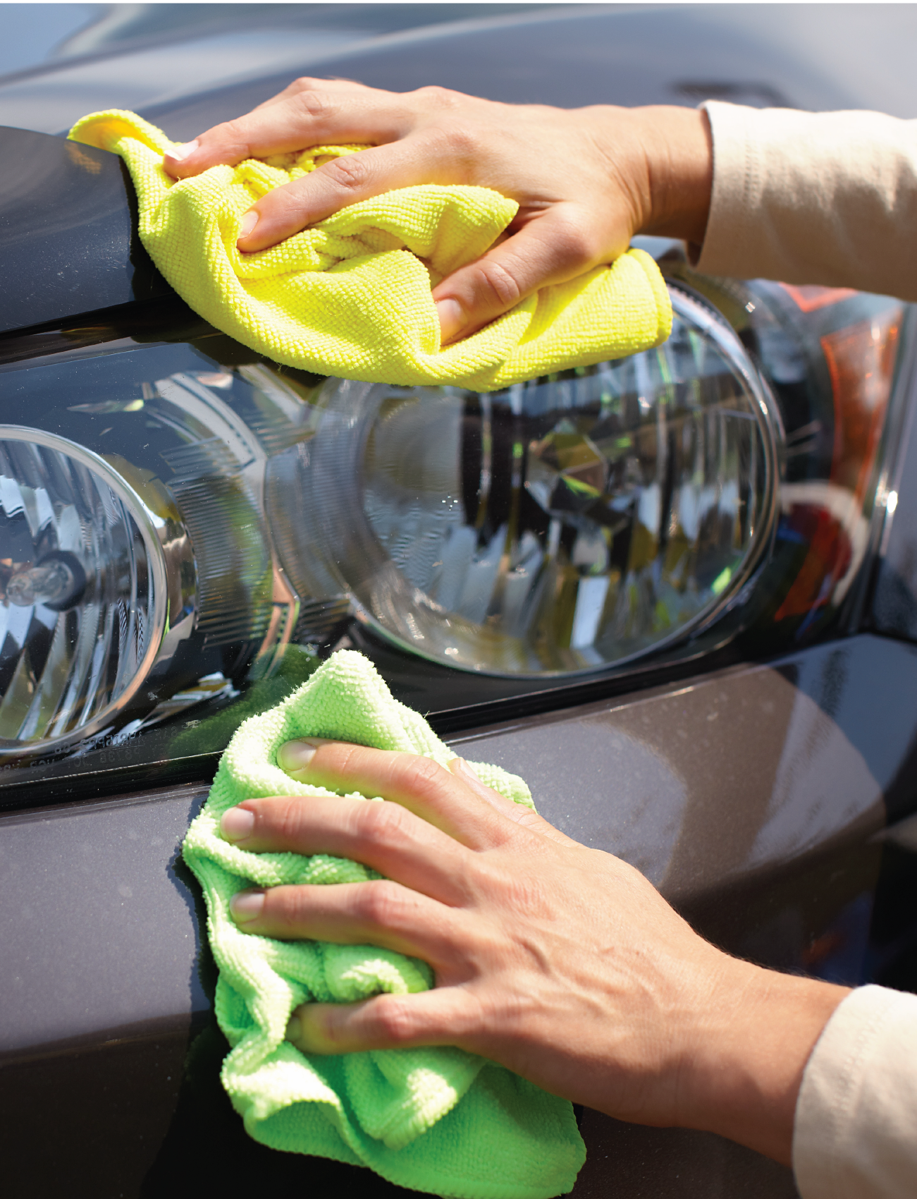 Two hands using Sunland-Protection's Ultra-soft Microfibre 6 x Towel Pack in yellow and green to clean and polish a car's headlight and hood, achieving a lint-free, streak-free shine in sunlight, indicating thorough detailing.