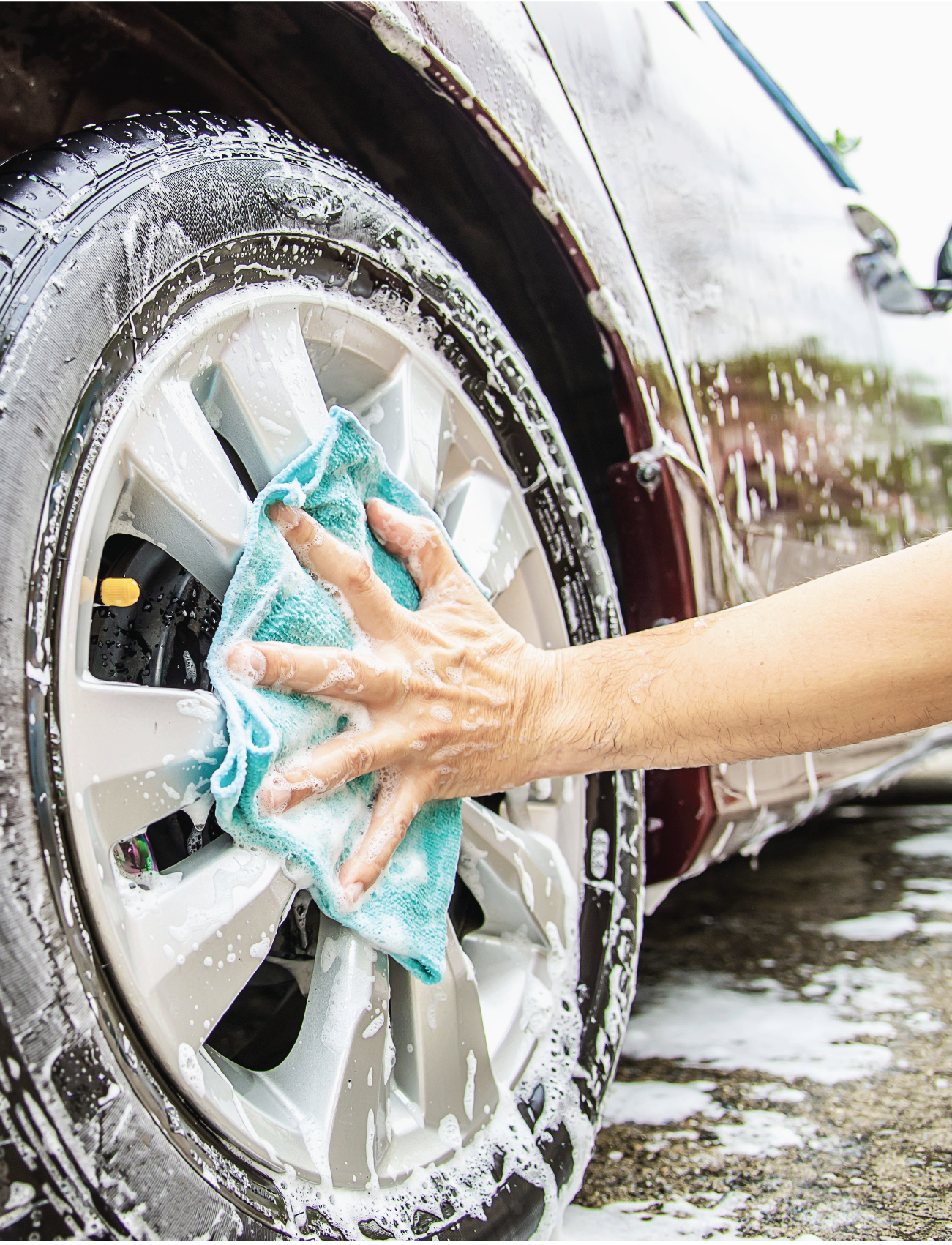 A person is cleaning a car tire with an Ultra-soft Microfibre towel from the Sunland-Protection 6 x Towel Pack, using soapy water. The high-density weave ensures a lint and streak-free finish. A close-up shows a shiny wheel with soap suds while the car stands on wet ground.