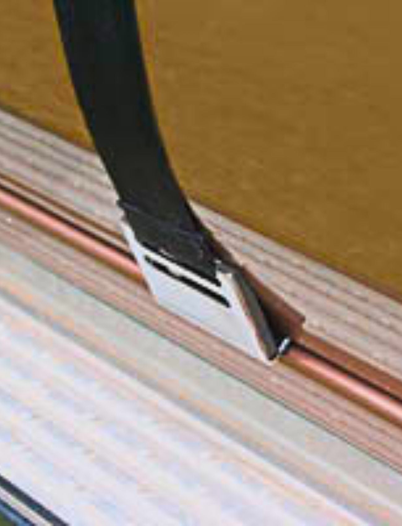 Close-up of a metal clamp securing a black strap onto a copper pipe, reminiscent of the sturdy design seen in Sunland-Protection's MIRROR WIDE DOOR BASE STRAP - SUITS MH3015. The clamp is fastened to a wooden surface, highlighting part of a layered board with varying shades.