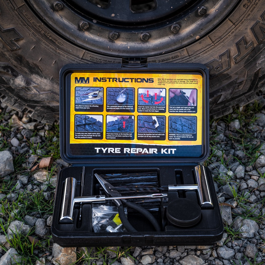 On a rocky ground in front of a vehicle tire, open the Mean Mother 4x4 Heavy Duty Tyre Repair Kit 28 Pce. Perfect for emergency situations, it offers everything needed for tubeless tyre repairs, complete with tools and materials, and features illustrated instructions on the case lid.