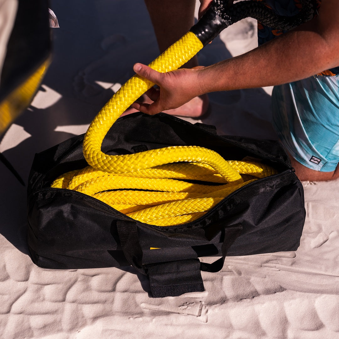 A person in blue patterned shorts kneels on sand, placing a Mean Mother 4x4 Kinetic Rope 9m - 9500kg into a black bag. The vibrant yellow rope suggests a beach activity, possibly testing its 30% stretch capability.