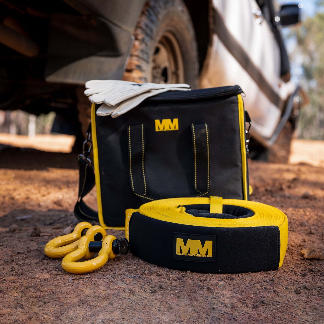 The Mean Mother 4x4 Recovery Kit, designed for 11,000 kg capacity and consisting of five pieces, is displayed on the ground. It includes a distinctive black and yellow bag, a heavy-duty snatch strap, bow shackles, and gloves. In the background, you can glimpse a muddy vehicle tire suggesting an exciting outdoor adventure setting.