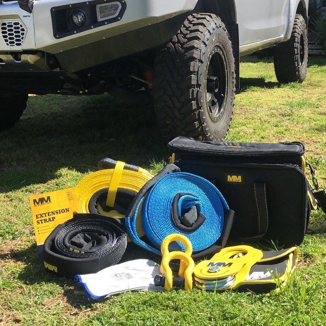 A Mean Mother 4x4 Recovery Kit 11,000kg 8pc | Premium is displayed on grass in front of a rugged vehicle, ideal for the dedicated 4WD enthusiast. This premium off-road recovery gear includes a yellow extension strap, blue and black tow straps, shackle hooks, gloves, and a black carrying bag.
